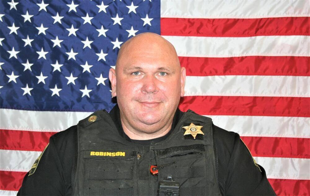 White male deputy in a black polo shirt with a gold badge on it sitting in front of the American Flag.