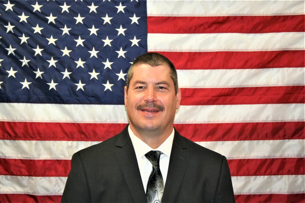 White male investigator in a dark dress suit with a red tie and white shirt sitting in front of the American Flag.