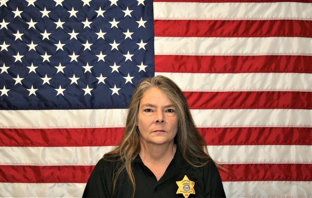 White female with black polo and gold badge sitting in front of the American Flag.