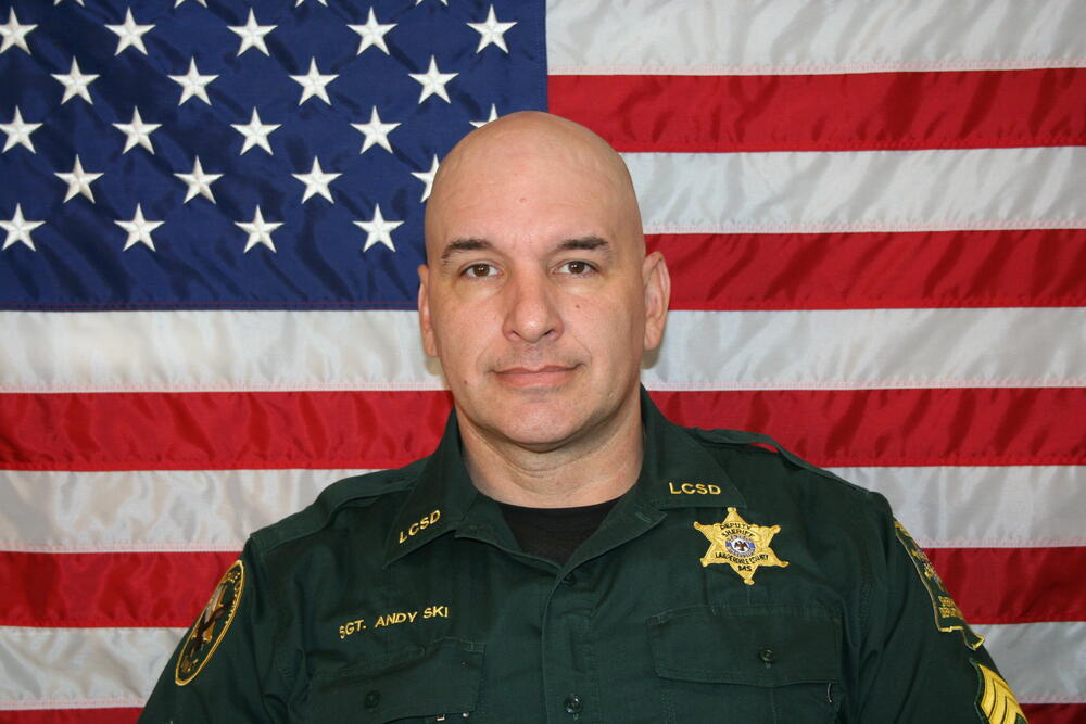 White male wearing a green LCSO uniform and sitting in front of the American Flag