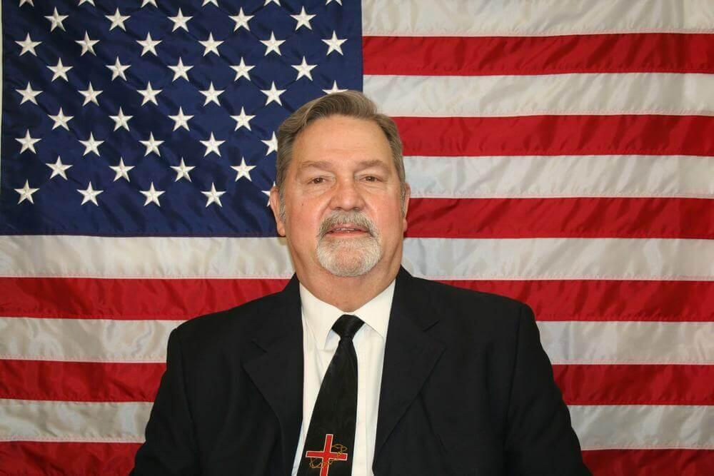 Dennis Marks standing in front of the flag of the United States of America.