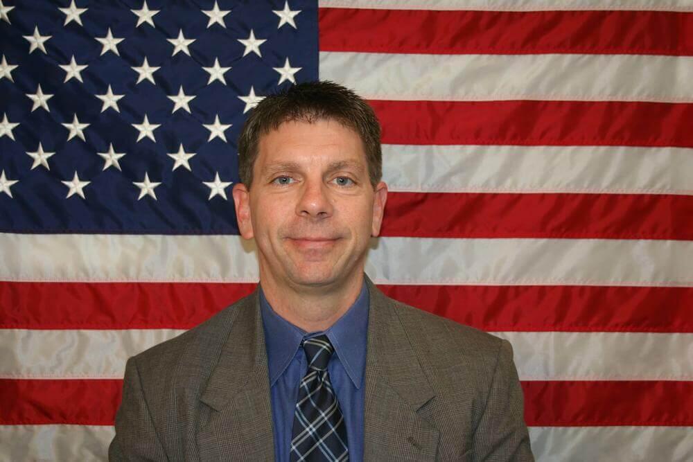 Major Greg Lea standing in front of the flag of the United States of America.