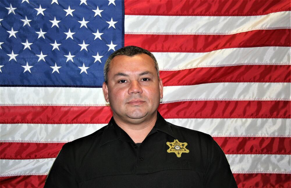 Major Jared Woodall in front of the American Flag wearing a black law enforcement uniform with a gold badge.