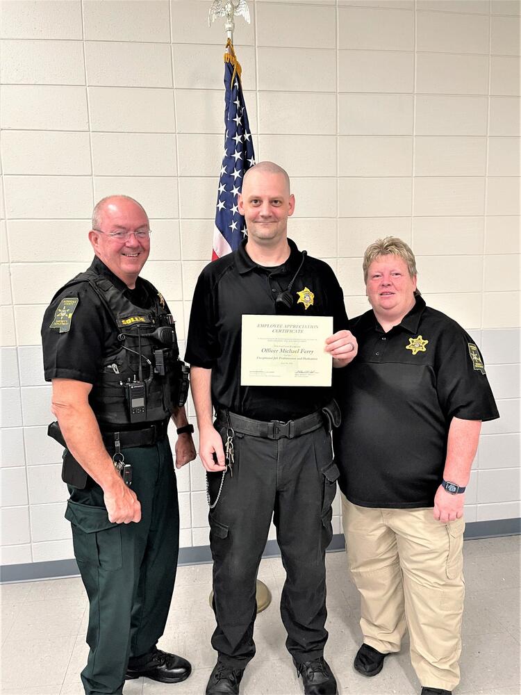Sheriff Billy Sollie/Detention Officer of the Month Michael Ferry/Jail Admin Melissa McCarter acknowleding holding certificate of recognition in front of the American Flag