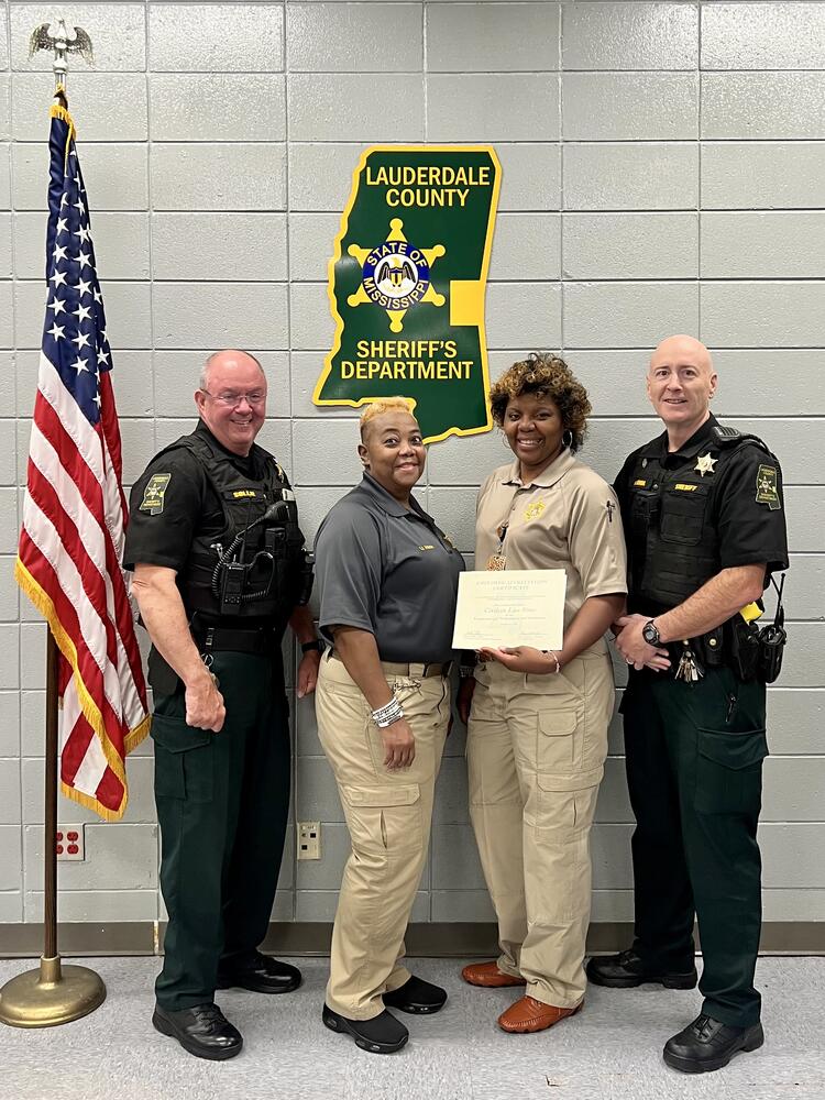 LCSO Sheriff, Chief, Lt. Maclin, Civilian Sim in front of green agency patch and American Flag holding certificate of appreciation