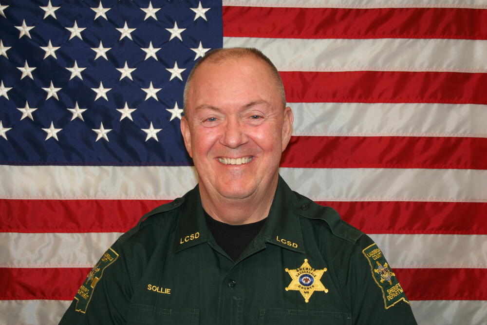 Sheriff Billy Sollie dressed in green patrol uniform in front of the American Flag