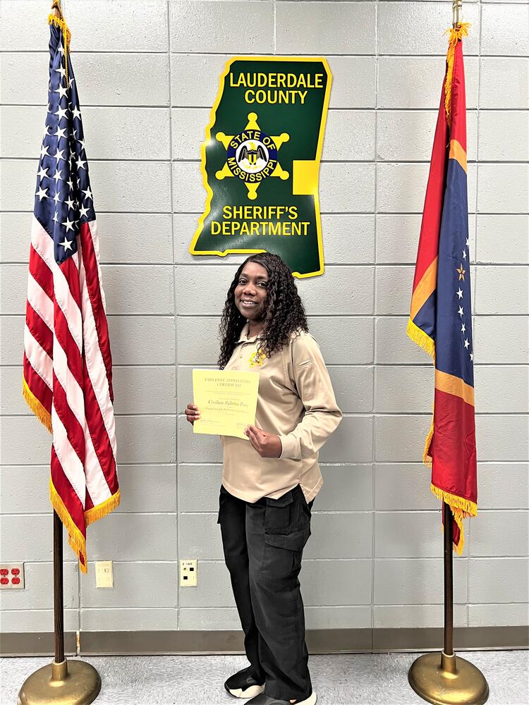 Black Female with khaki oxford shirt and gold jail badge wearing black pants. Red, white and blue Magnolia Mississippi flag and American Flag on each side. She is holding a gold certificate.