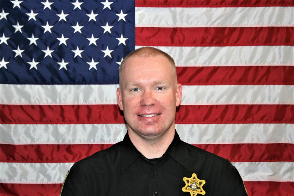 White male deputy with black polo shirt and gold badge in front ot the American Flag