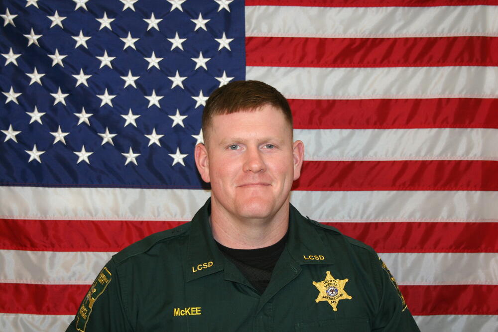 Sgt McKee in a green law enforcement uniform with gold badge in front of the American Flag
