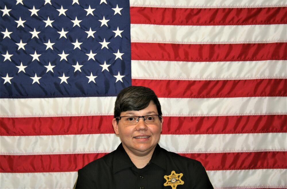 White female deputy with a black polo shirt and a gold badge sitting in front of the American Flag.