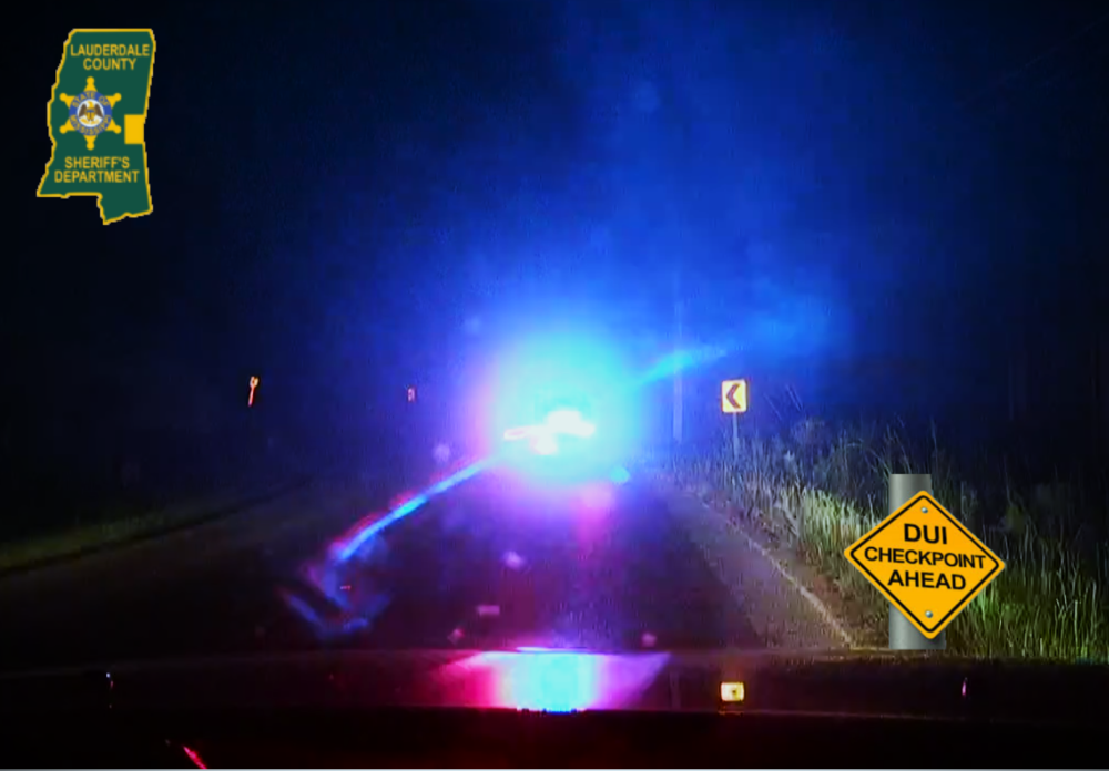Looking through a vehicle windshield at night and seeing a yellow sign with DUI Checkpoint on it and a police car with blue lights ahead. Also, a green and yellow state of MS Lauderdale County Sheriff's Office logo on the upper left.