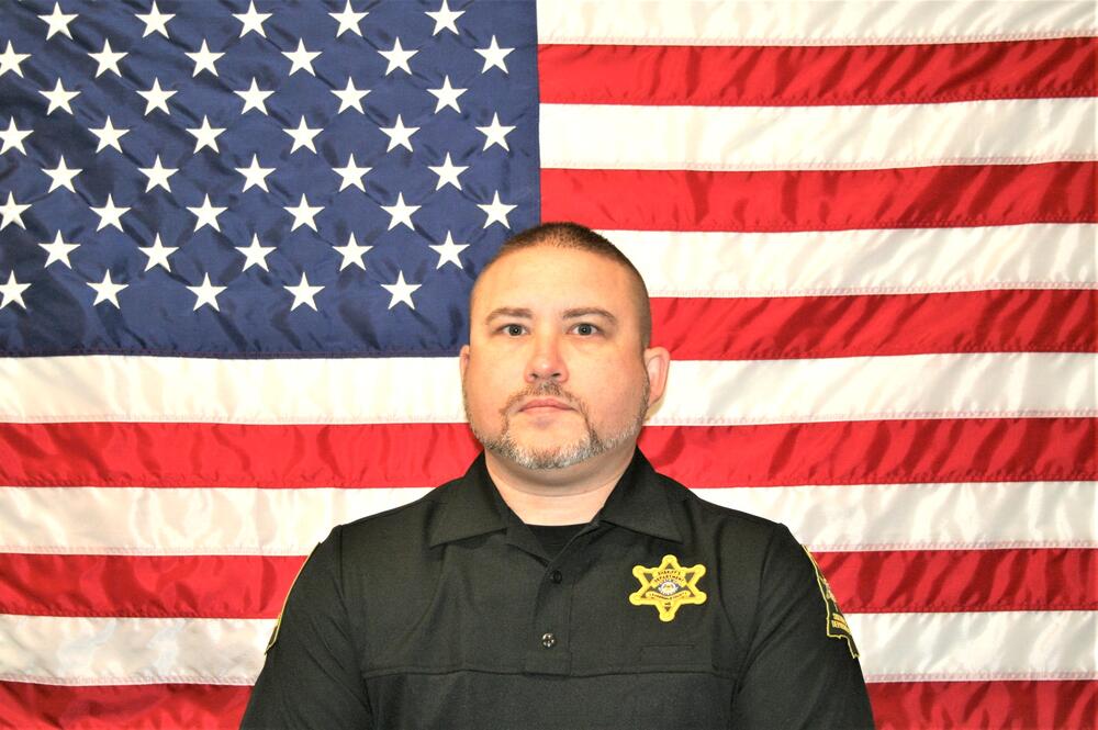 White male deputy in black polo with a gold badge sitting in front of an American flag.