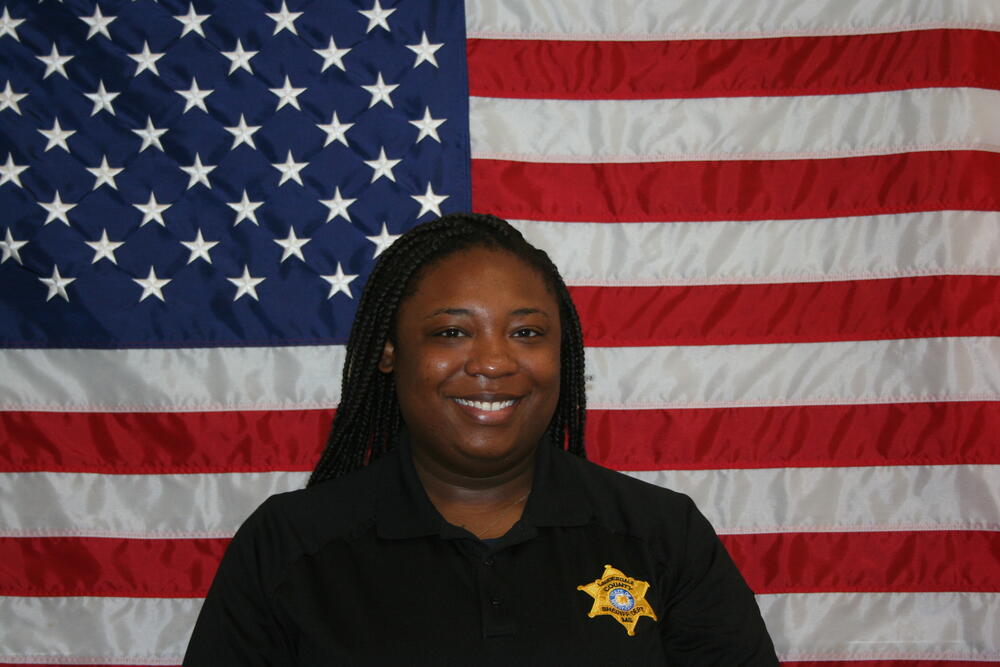 Black female with black polo and gold badge in front of the American Flag