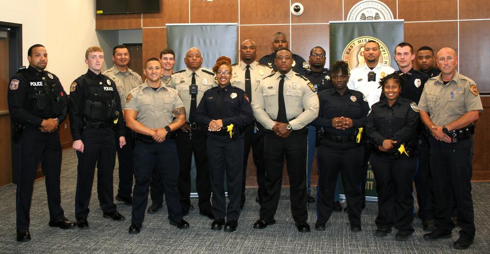 Multi-Race Police Officers in uniform standing in front of green Crisis Intervention Team banners hold graduation certifications from the week's class.