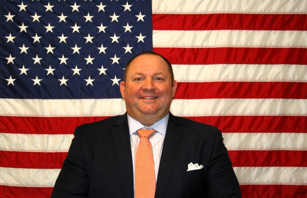 Major Toby Pinson sitting in front of the American Flag in a blue suit with orange tie.