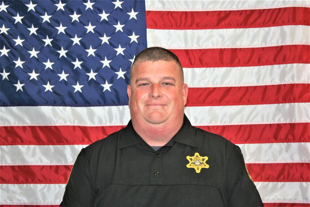 Brian Fortenberry sitting front of the American Flag wearing a black uniform shirt with a gold badge.
