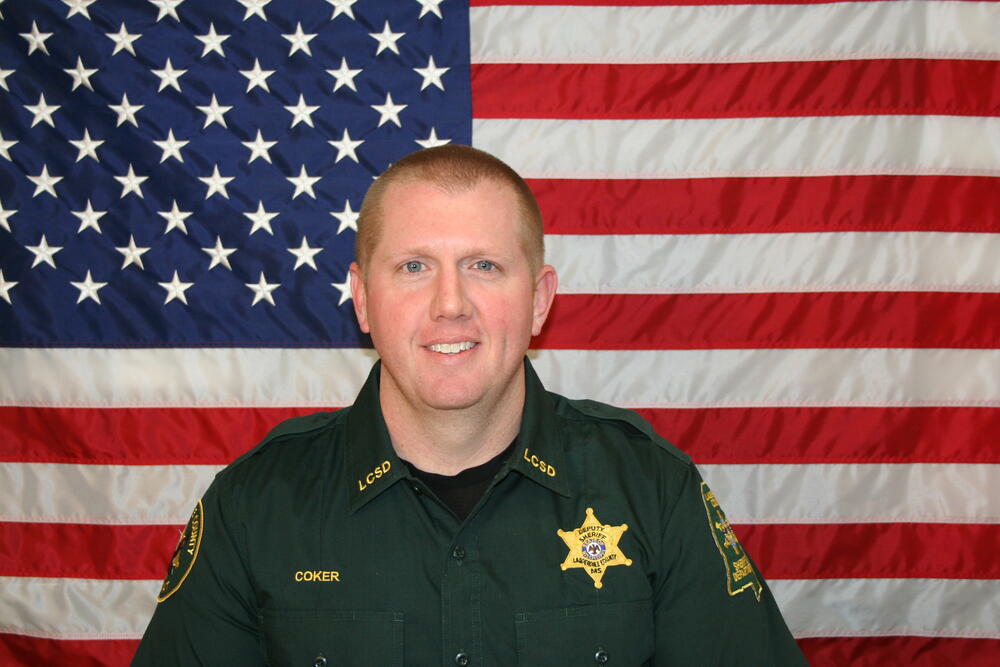White male in a green sheriff's office uniform in front of the American Flag.