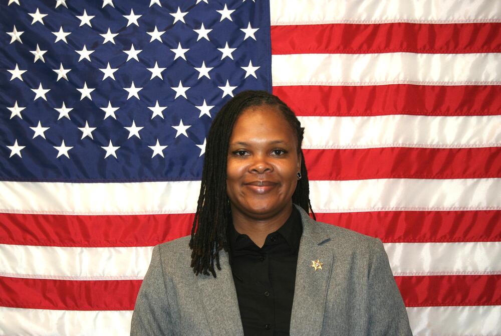 Black female investigator wearing a gray suit and black blouse in front of the American Flag.