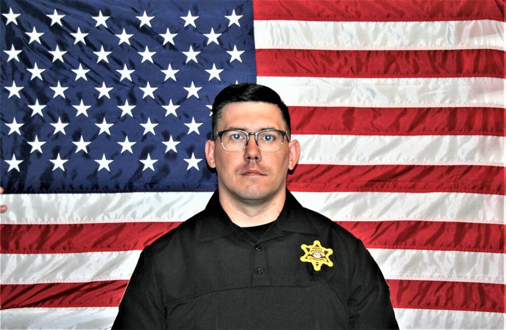 White male School Resource Officer with black polo and gold law enforcement badge sitting in front of the American Flag