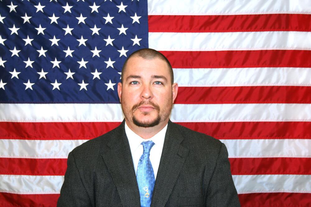 White male, sandy brown hair and mushtache wearing a dark grey suit coat and blue tie.