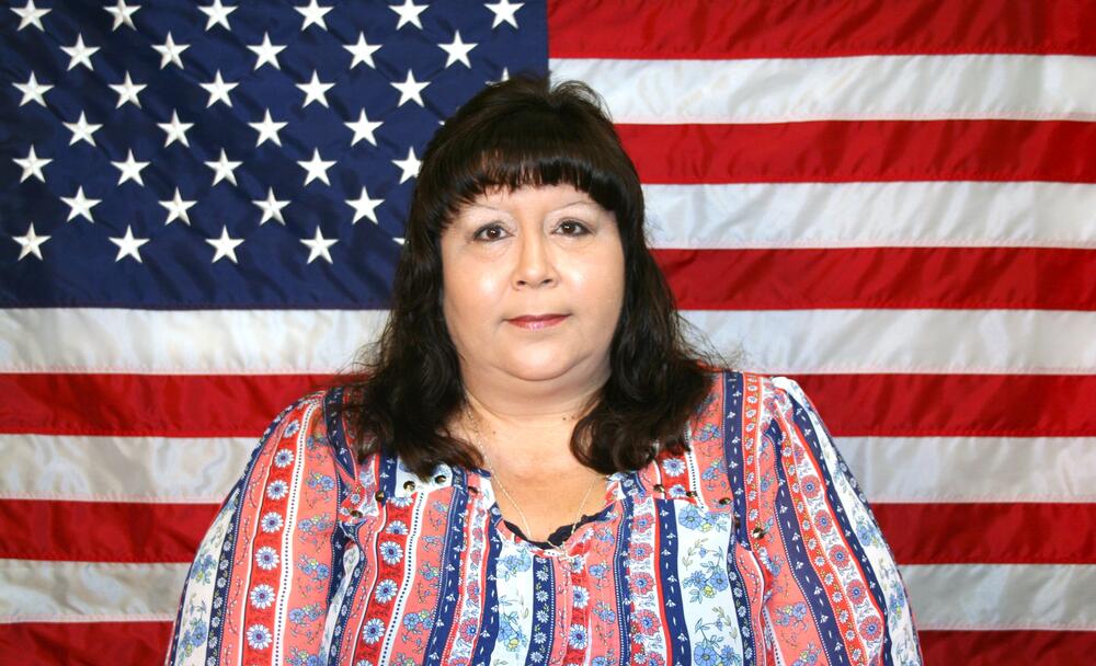 White female with long dark hair wearing a multicolor blouse.