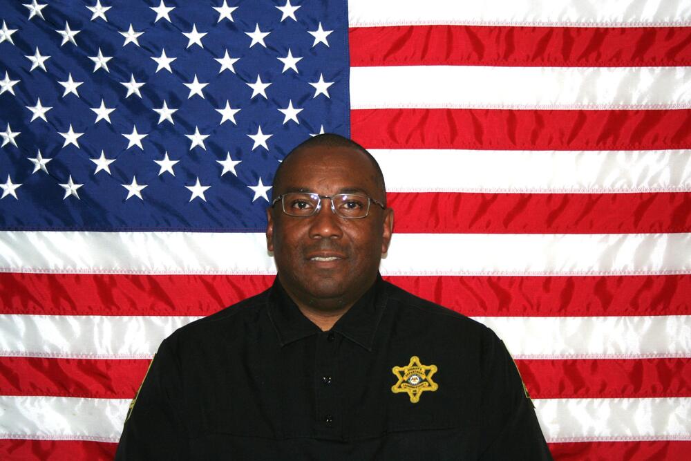 Black male wearing glasses sitting in front of the American Flag wearing a black polo and gold badge