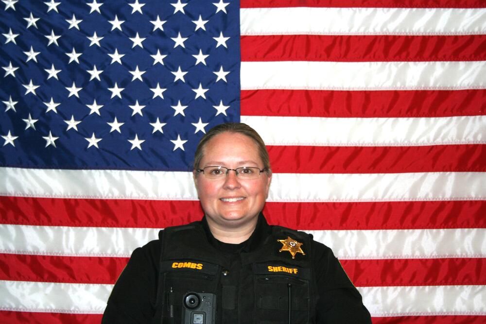 White female wearing glasses and sandy hair in a bun sitting in front of the American Flag. Marissa is wearing a black bulletproof vest with a gold sheriff's badge on it.