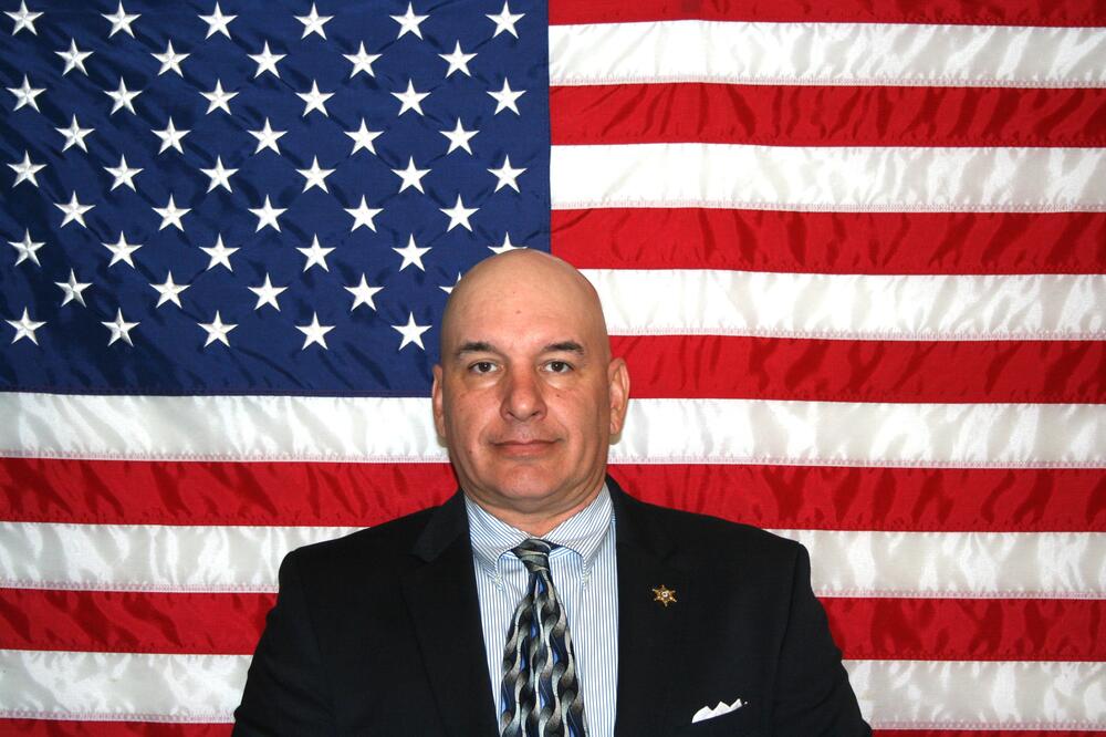 White bald male sitting in front of the American Flag wearing a dark suitcoat and multicolored tie.