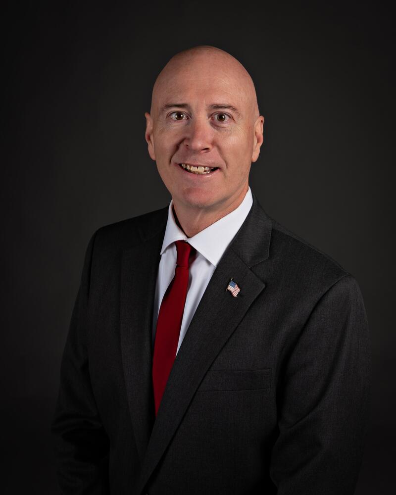 White male with bald head and dark suit with red tie.