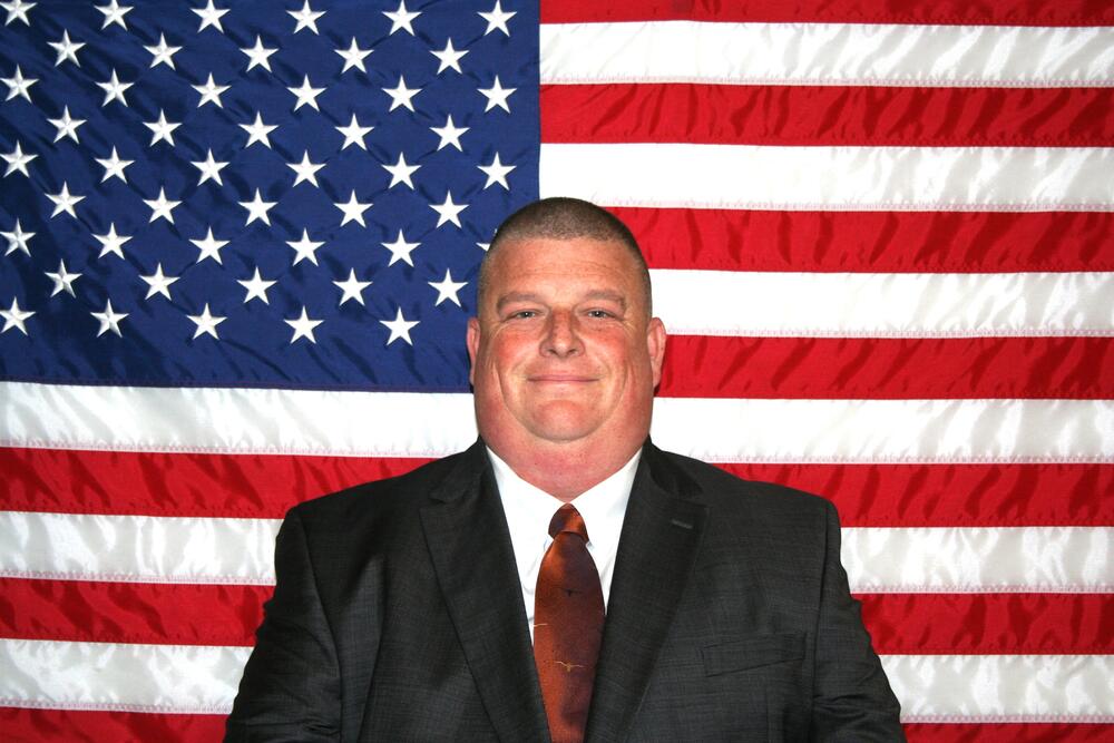 White male with dark hair wearing a dark suit coat with a Texas Longhorn tie.