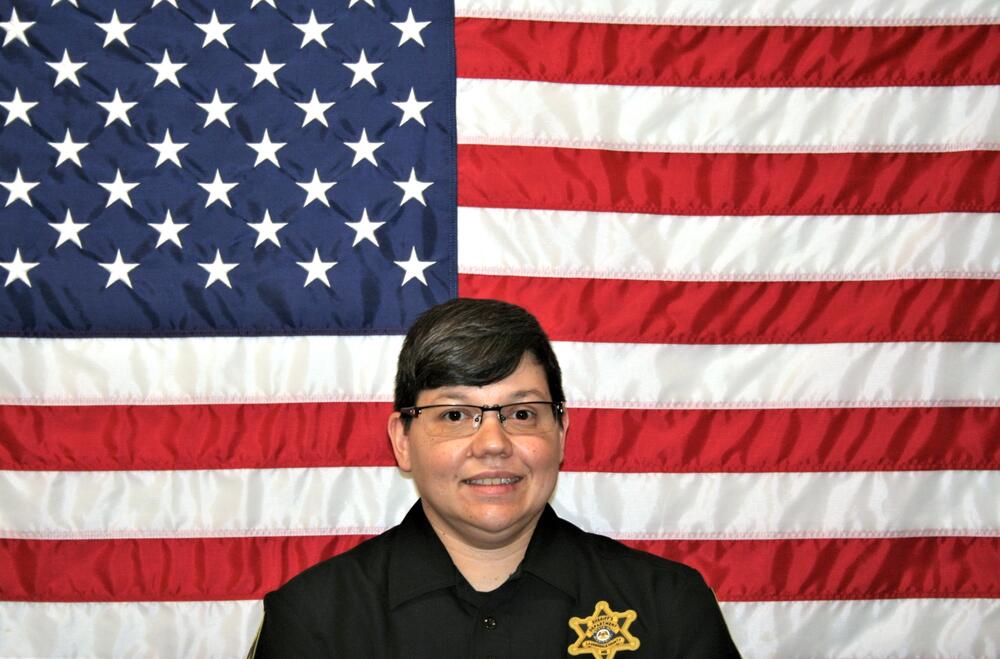 White female with dark hair wearing a black Sheriff's Office polo in front of the American Flag.