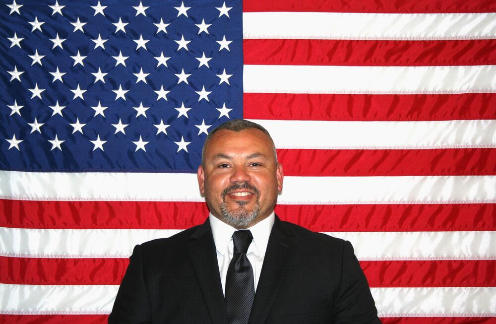 White male with salt and pepper hair wearing a dark gray suit and tie sitting in front of the American Flag.