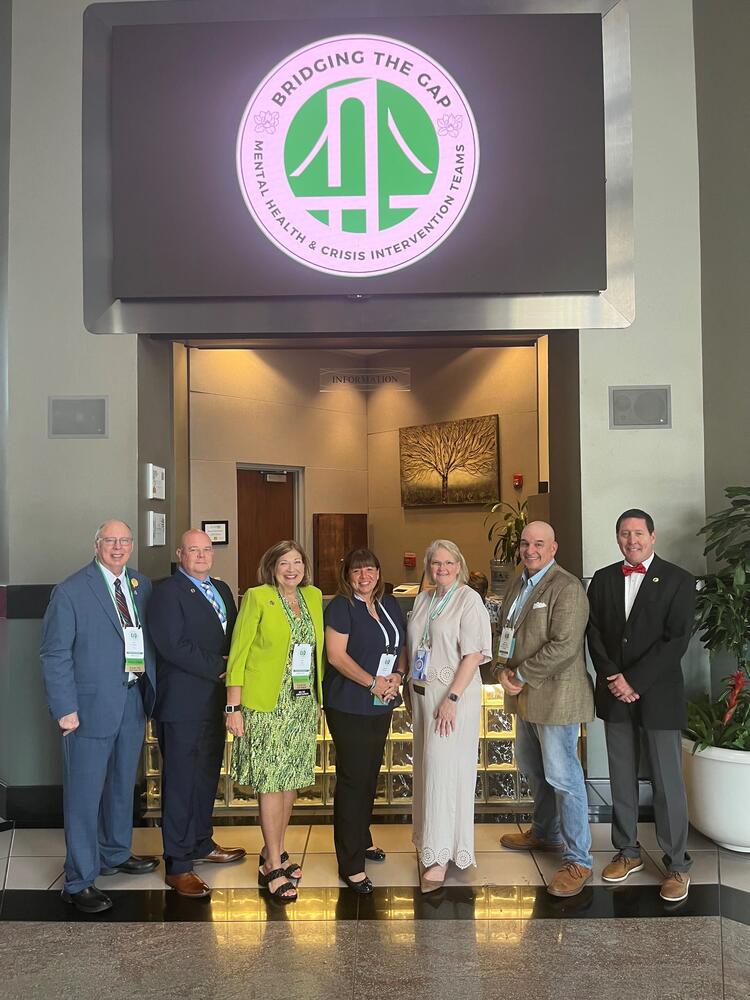 3 White females and 4 White males wearing dress attire standing in front of an office entrance with brown tree painting on the wall.