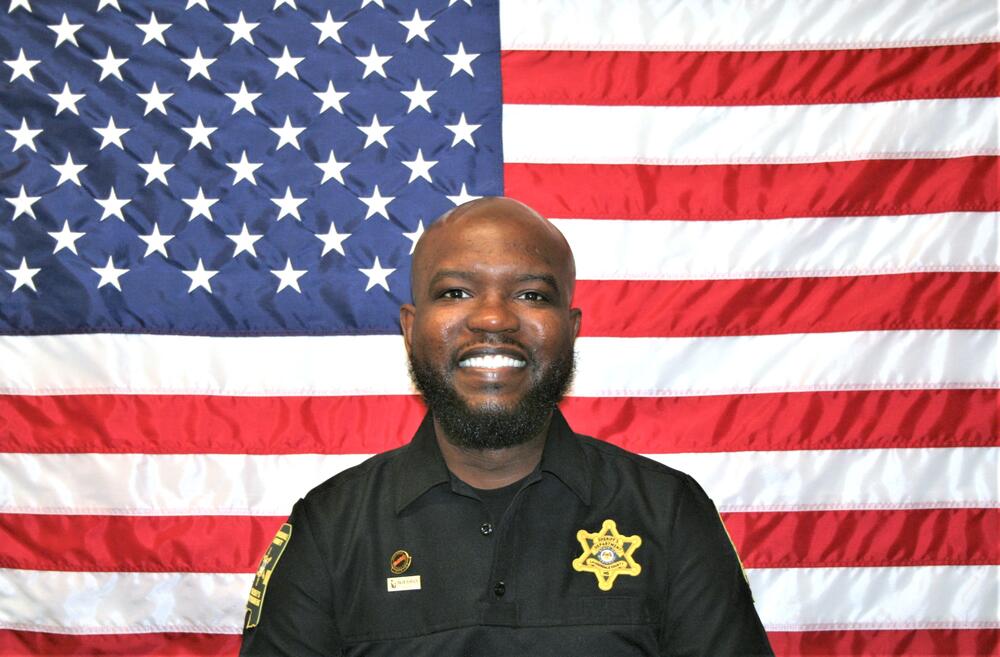 Black male sitting in front of the American Flag wearing a black uniform shirt with a gold sheriff's badge on it.