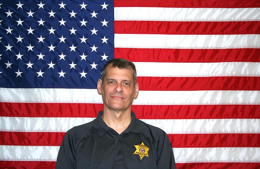 White male with sandy short hair wearing a dark gray polo with a gold sheriff's badge.